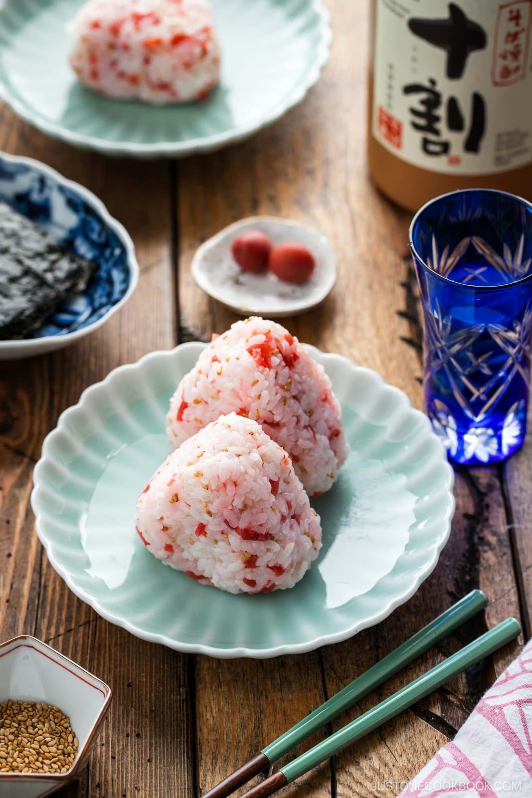 Two pink Umeboshi Onigiri (Plum Rice Ball) from Midnight Diner sit on a scalloped plate, surrounded by nori, umeboshi, a glass of water, sesame seeds, and a bottle, all arranged on a wooden table with chopsticks nearby.