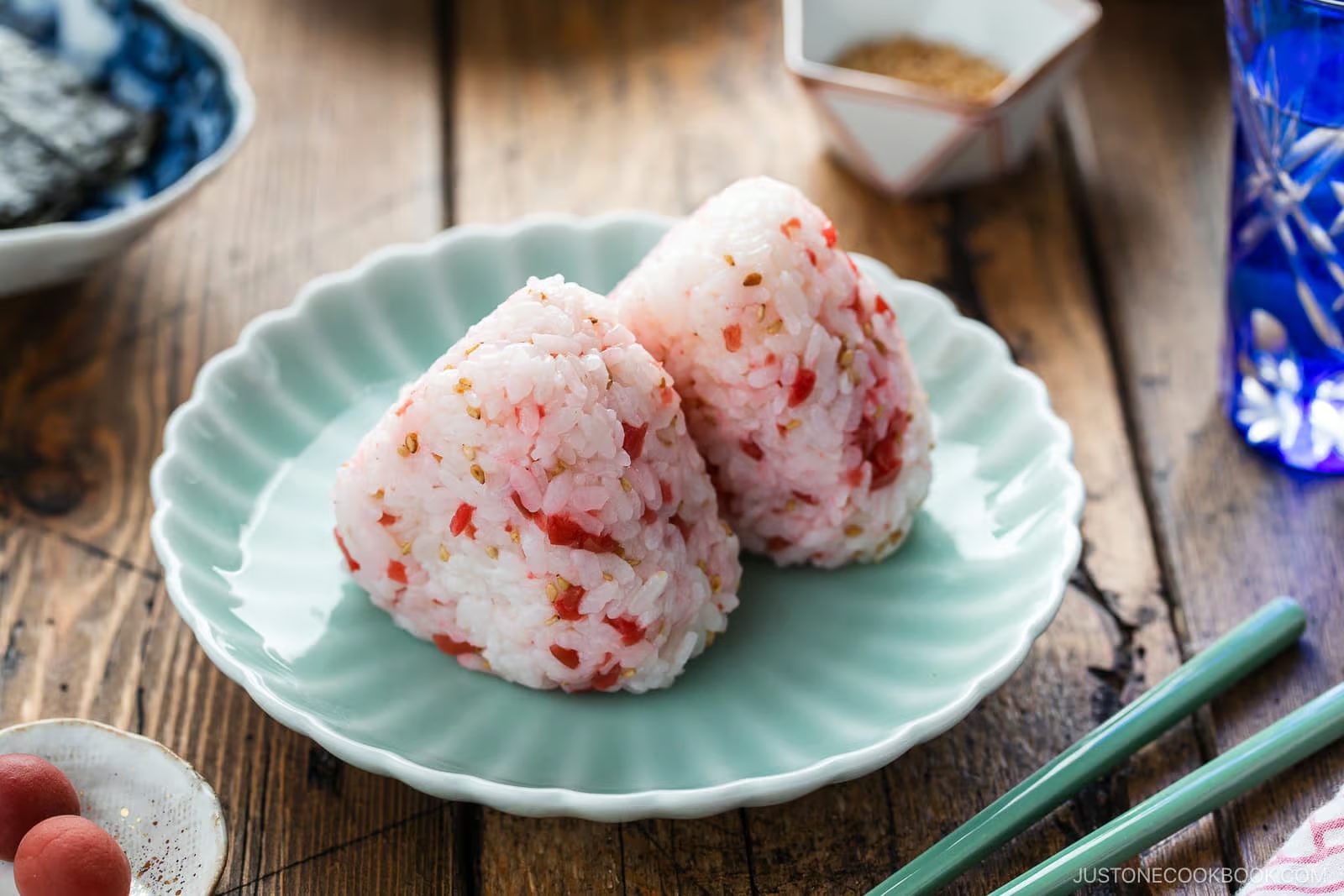 Two Umeboshi Onigiri (Plum Rice Balls), inspired by Midnight Diner, with red seasoning sit on a pale-green plate, accompanied by green chopsticks. A wooden table, pickled garnish, water glass, and small dishes are visible in the background.