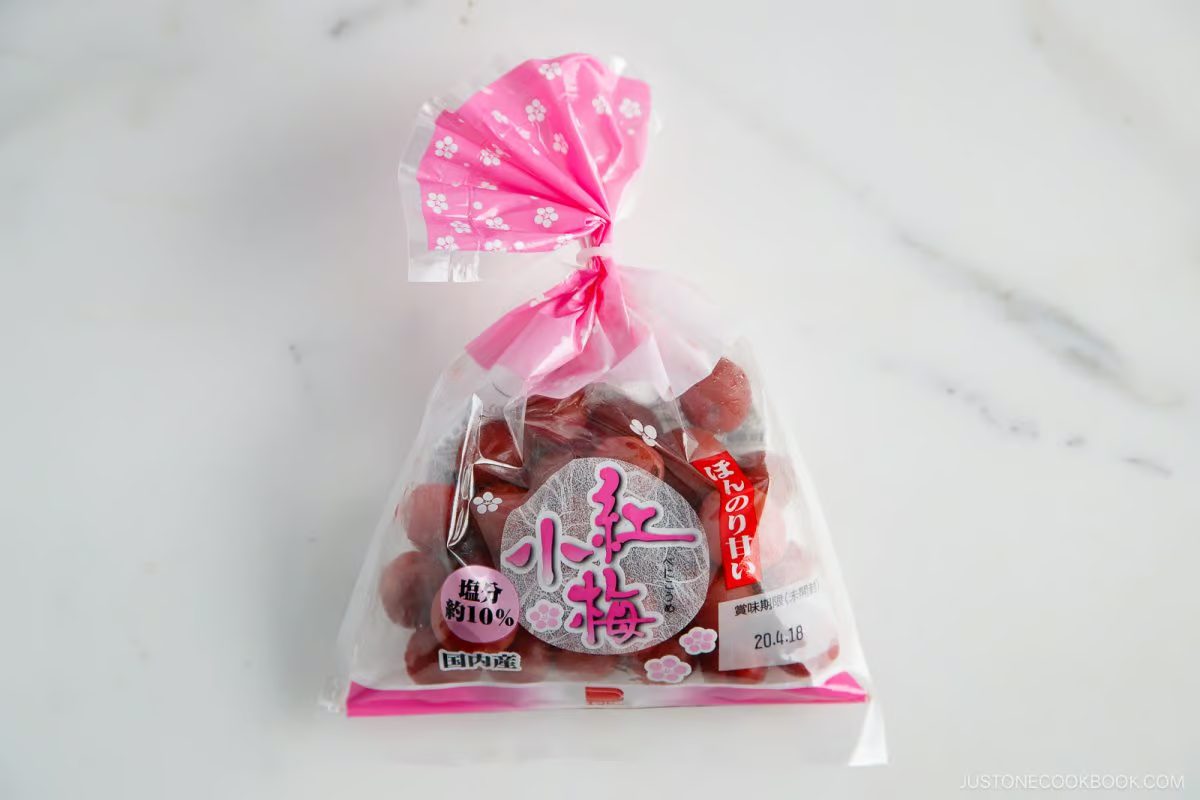 A clear plastic bag with a pink floral design contains red candied umeboshi (Japanese salted plums), often enjoyed in Umeboshi Onigiri (Plum Rice Ball) as seen in Midnight Diner. The bag has Japanese writing and nutrition details visible on a white marble surface.